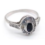 An 18 K white gold ring with a central dark blue sapphire and diamonds on the halo and shoulders.