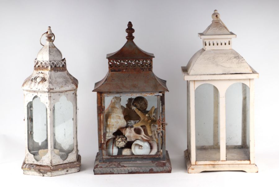 Three painted metal storm lanterns, one containing a collection of seashells, largest 53cms high (