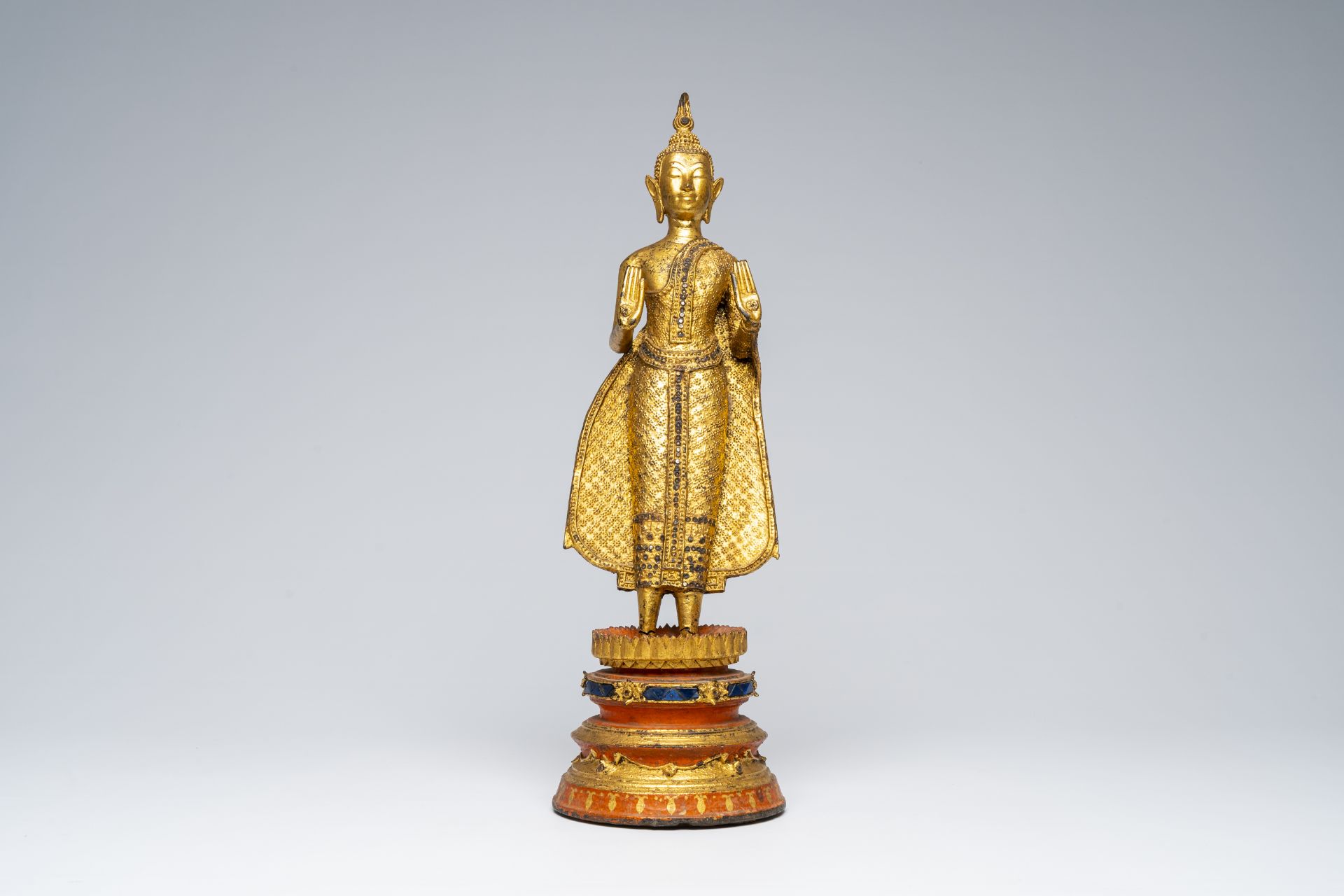 A Thai gilt bronze figure of a standing Buddha on a polychrome painted and glass inlaid metal base,