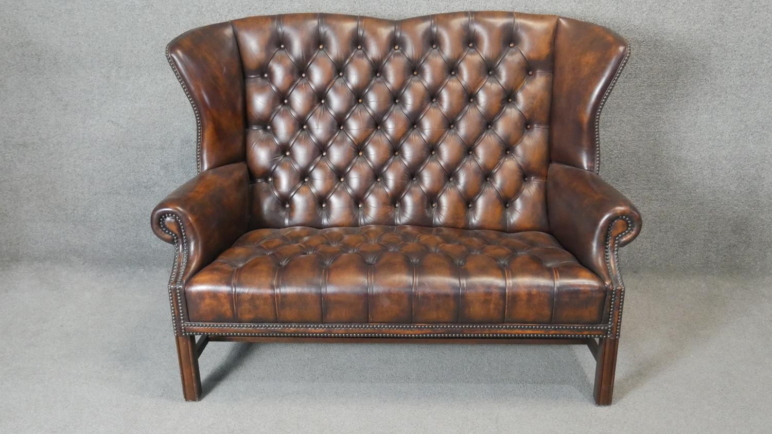 A Georgian style wingback two seater armchair in deep buttoned and studded leather upholstery on