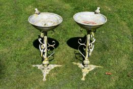 A pair of distressed painted birdbaths with a bird perched on their rims raised on scrolling wrought