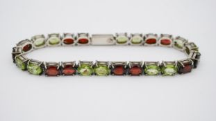 A silver peridot and garnet set line bracelet. Set with twelve oval mixed cut peridots in open