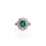 An Edwardian emerald and diamond cluster ring; the stepped rounded rectangular emerald in