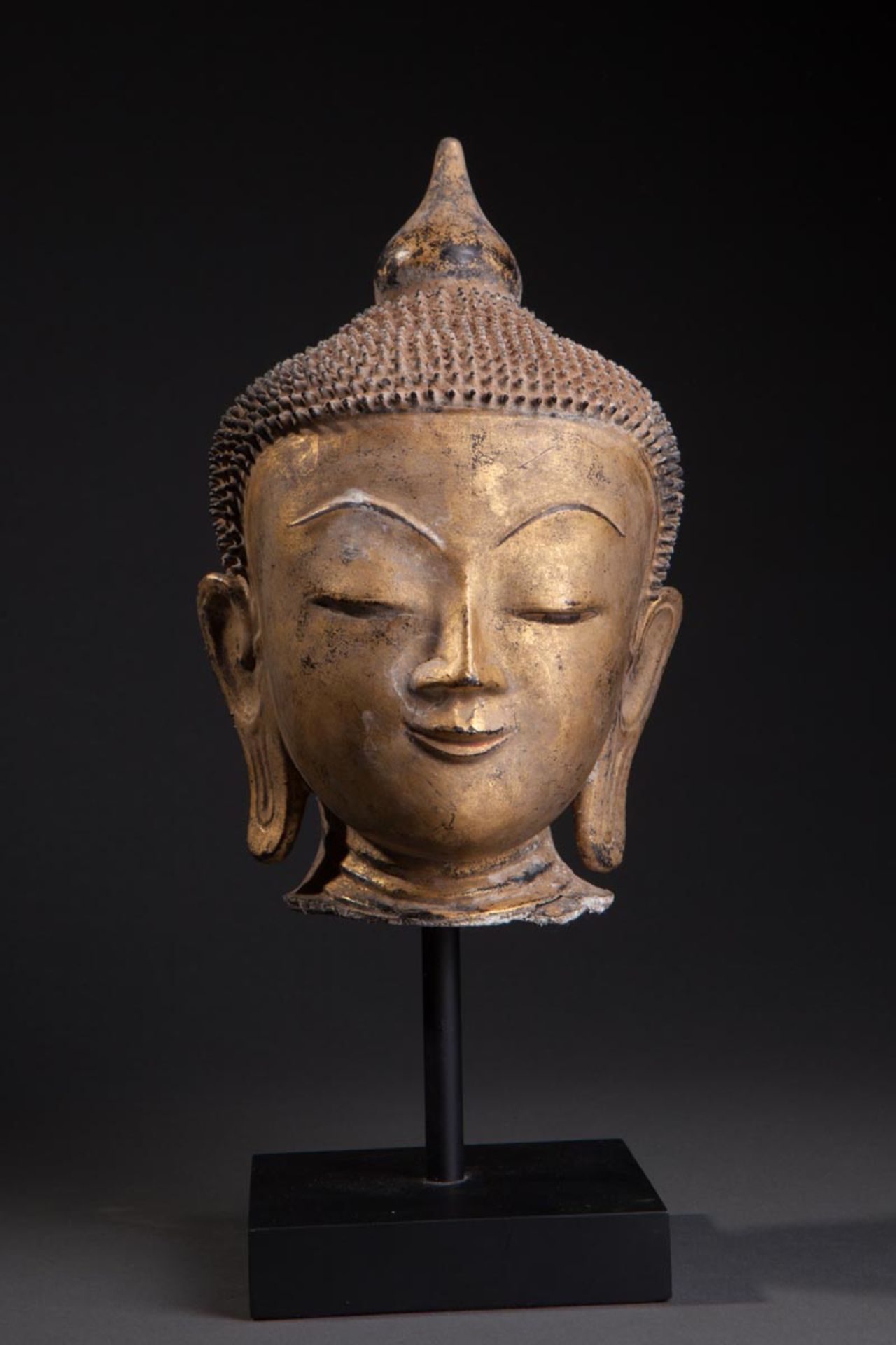 Tête de Buddha à l'expression sereine, la coiffure hérissée de bouclettes surmontée d'un rasmi
