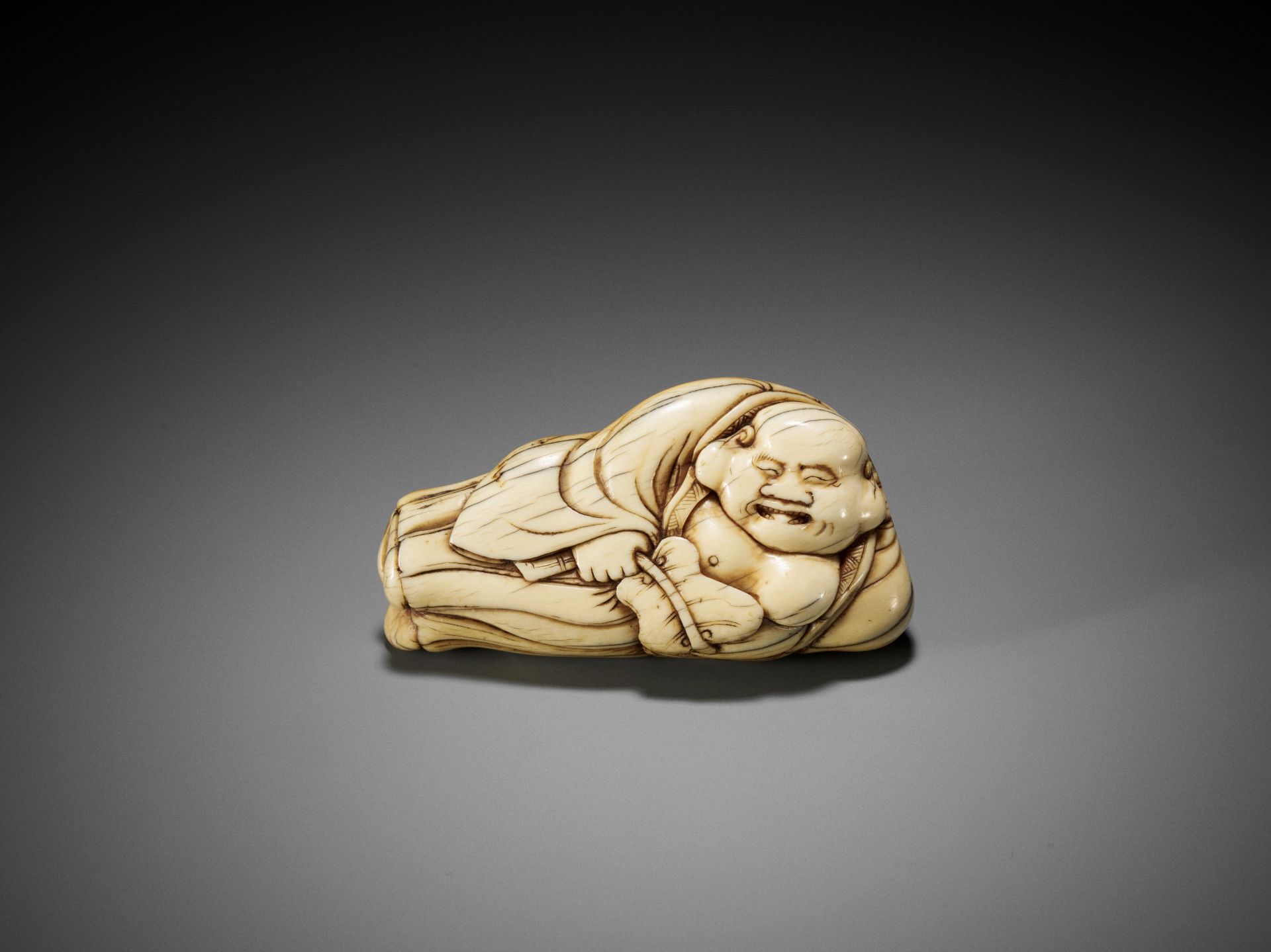 AN IVORY NETSUKE OF A RECLINING HOTEI WITH UCHIWA (FAN)