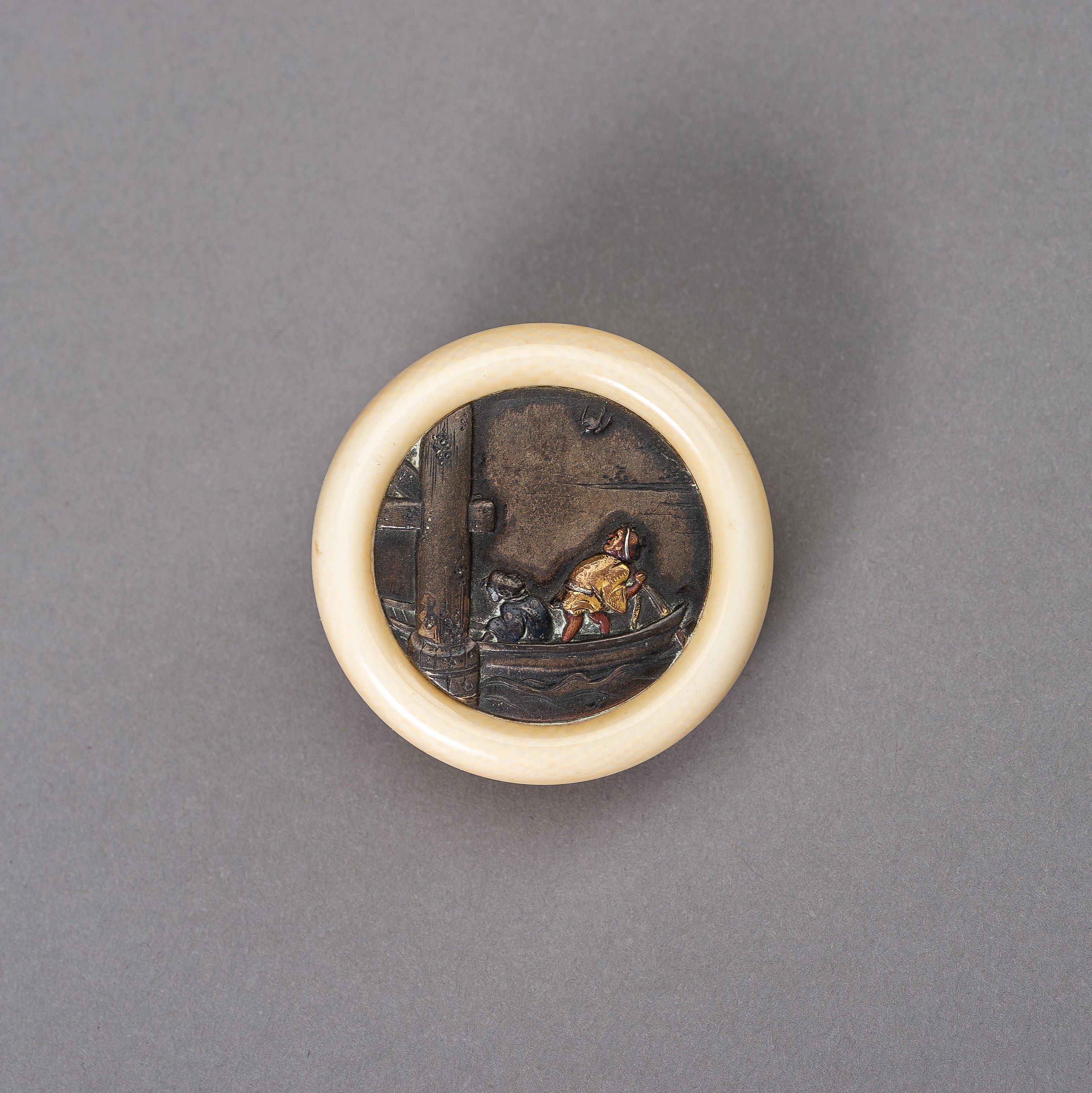 AN IVORY AND MIXED METAL KAGAMIBUTA NETSUKE DEPICTING TWO FIGURES ON A BOAT