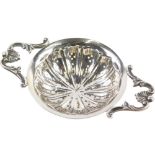 A George V silver tea strainer, the circular body flanked by scroll orb and shell handles, with pier