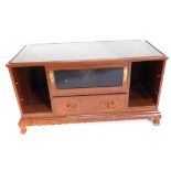 An Eastern hardwood television table, the panelled top with a glass inset above a door and a carved