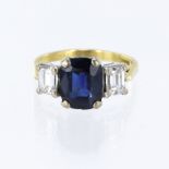 18ct yellow gold trilogy ring, set with a cushion cut mid-blue sapphire measuring 10mm x 8mm total
