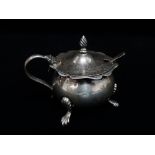 An Edwardian silver hinge lidded mustard pot with blue glass liner, raised on three outswept feet,