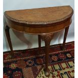 A Queen Anne style demi lune walnut card table with figured and carved top opening to reveal a baize