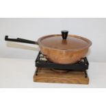 A vintage copper saucepan & burner on a metal trivet with a wooden base.