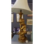Modern gilded finish table lamp in the form of a seated cherub on scroll decorated base, with shade.