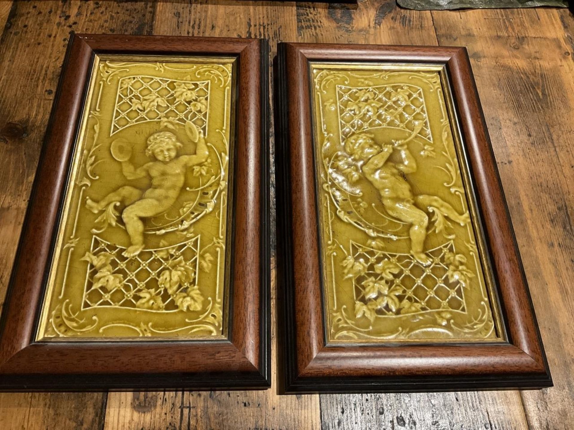 A Pair Of Framed Painted And Glazed  Tiles Depicting Putti / Cherubs  Playing Musical Instruments In