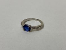 A white metal ring with single oval cut blue stone flanked by five diamonds to each shoulder, size