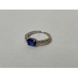 A white metal ring with single oval cut blue stone flanked by five diamonds to each shoulder, size