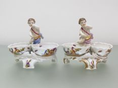 A pair of Berlin porcelain figural salts, each modelled with a putto and twin oval ozier-moulded