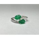 A two stone emerald ring on diamond-set shoulders, the oval-cut stones claw-set in a crossover