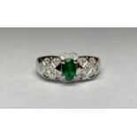 An emerald and diamond cluster ring, the central oval-cut emerald claw-set on a shaped plaque of