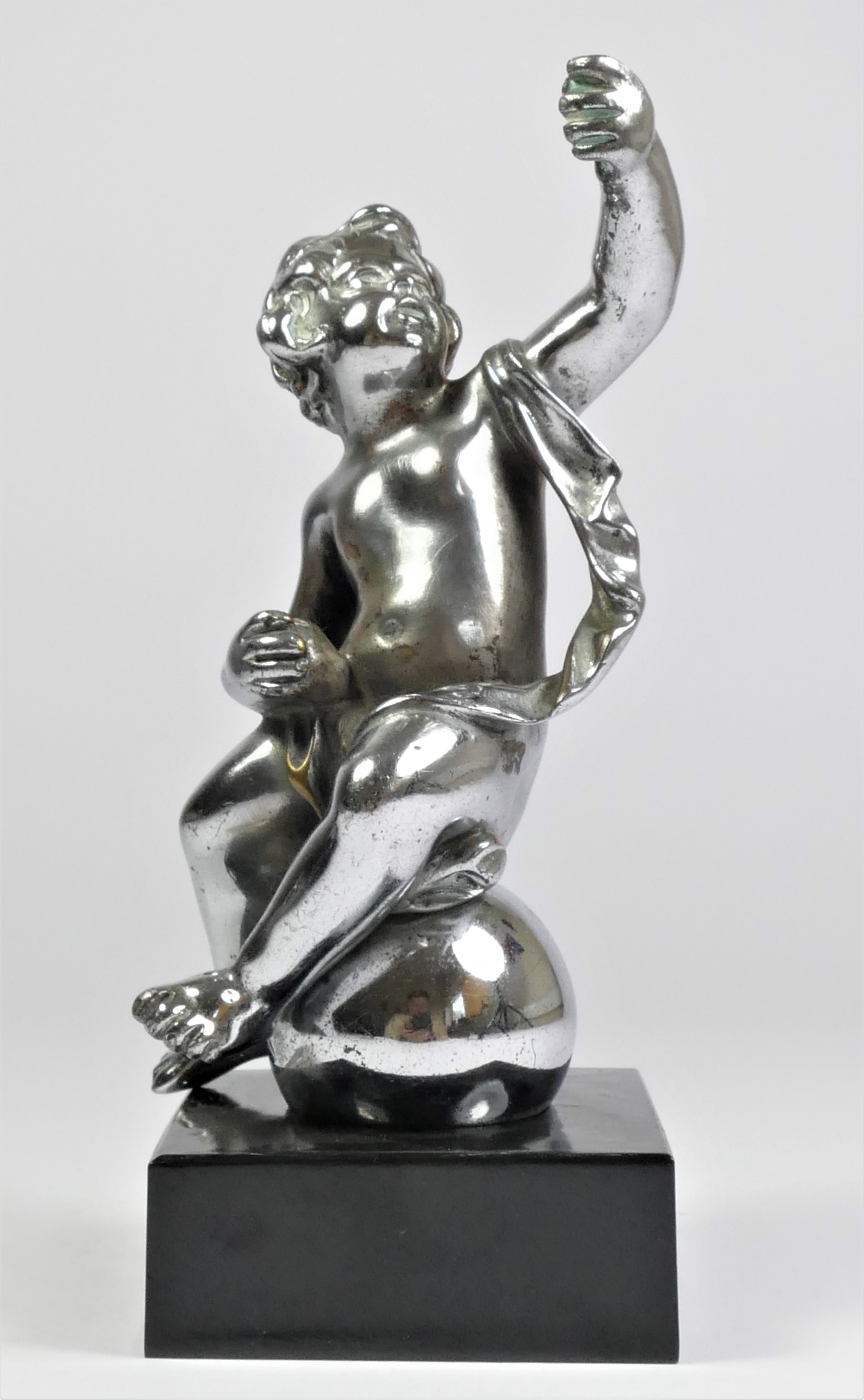 A chrome car mascot in the form of a cherub seated on a ball, mounted on a black onyx base, 18cm.