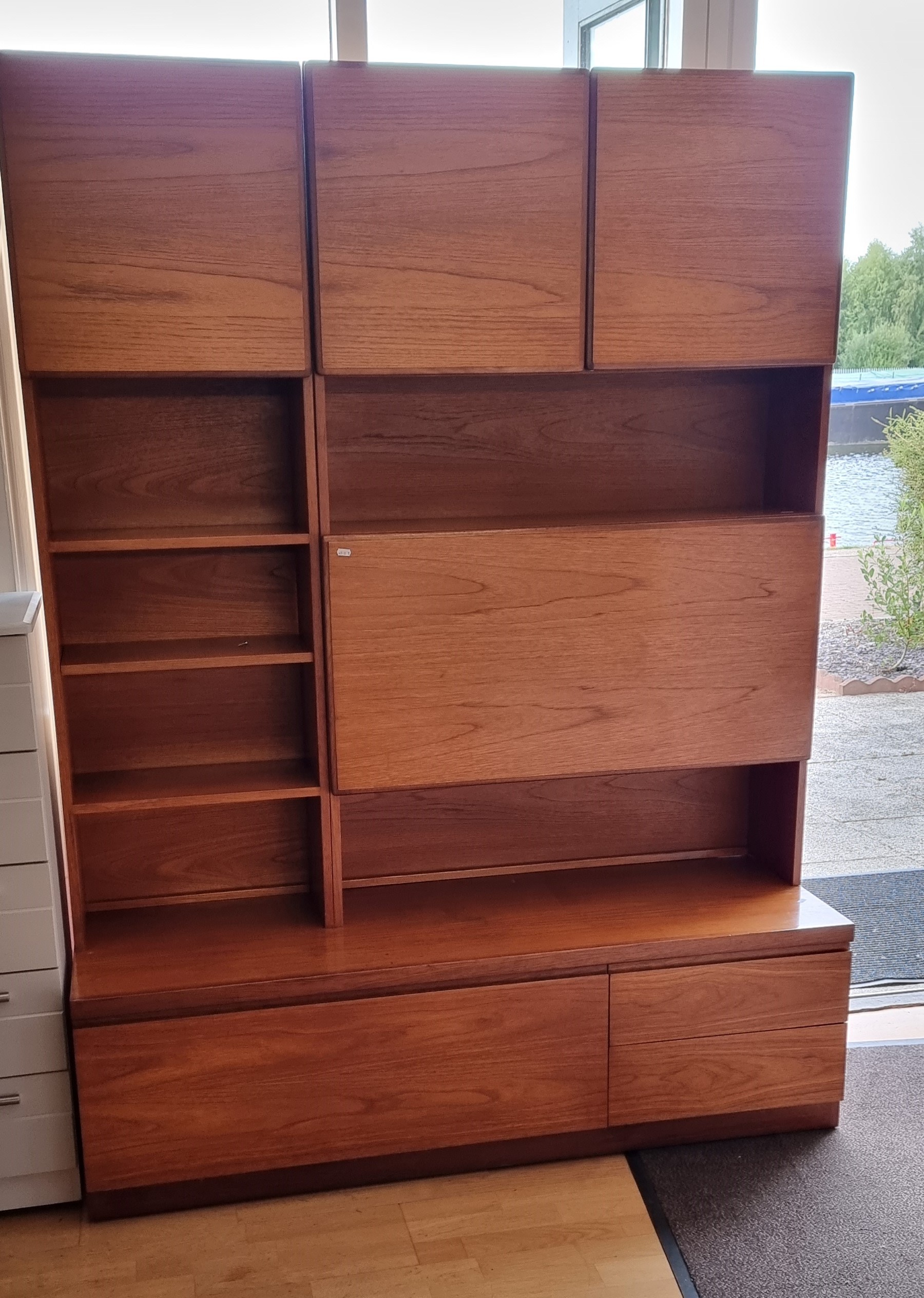 A teak display unit with three cupboard doors over shelving with drop down drinks cabinet over two