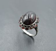 An oval garnet cabochon ring with garnet chip surround in a white metal collared mount on white