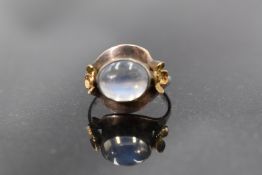 A moonstone cabouchon ring in a white metal collared mount flanked by two yellow metal flowers on