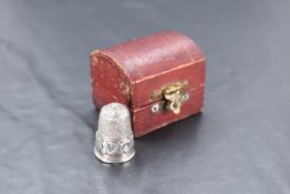 A white metal thimble, of traditional design, decorated with a band of scrolls centred by a shield-