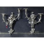 A pair of silvered metal twin branch wall sconces each with a pair of cherubs to the centre, 9” wide