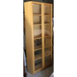 A light oak-finish tall standing cabinet fitted five shelves enclosed by a pair of glazed doors,