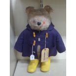 A soft toy Paddington Bear, wearing a duffle coat and yellow Wellington boots  bears a label  20"h