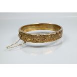 A silver-gilt half-hinged oval bangle with foliate engraved decoration, and safety chain attached,