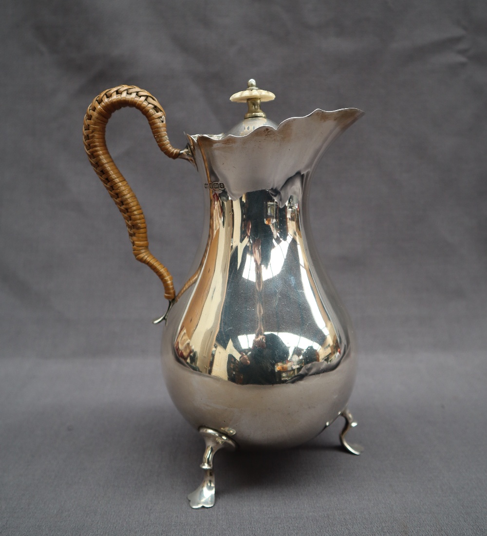 An Edward VII silver hot water jug with a flared edge and baluster body on three legs with trefid