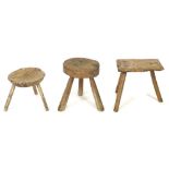 A group of three milking stools, two circular and one rectangular, all with splayed legs, tallest