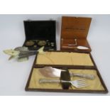 A set of silver plated fish servers, a cased foie gras set, a cased set of brass balance scales
