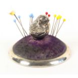A silver pin cushion, the circular base with domed velvet infill centred by a silver figure of a