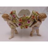 A continental porcelain table centrepiece in the form of three putti supporting a floral dish, 24