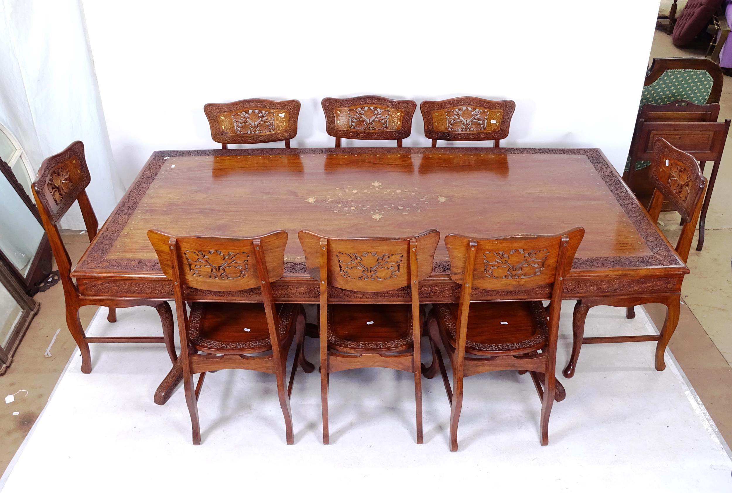 An Indian hardwood dining room table and 8 chairs, the table having floral carved edge with brass