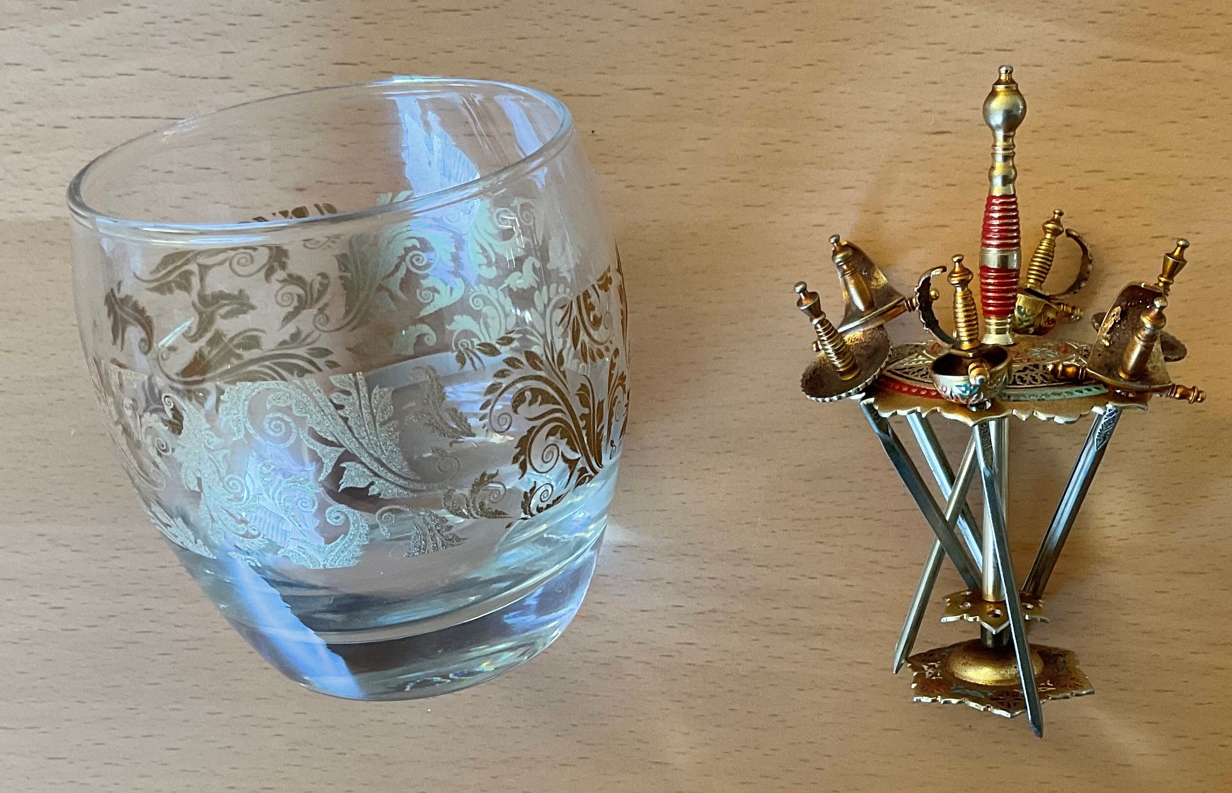 A small tumbler glass with 6 miniature gold plated swords. Glass measures 3.5 inches in height.