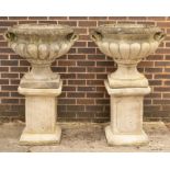 A pair of composition stone urns of lobed form with twin handles, on square plinths,
