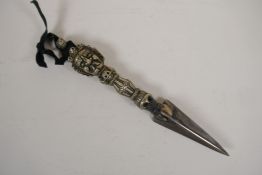 A Tibetan white metal phurba, the handle decorated with a wrathful deity mask and skulls, 9" long