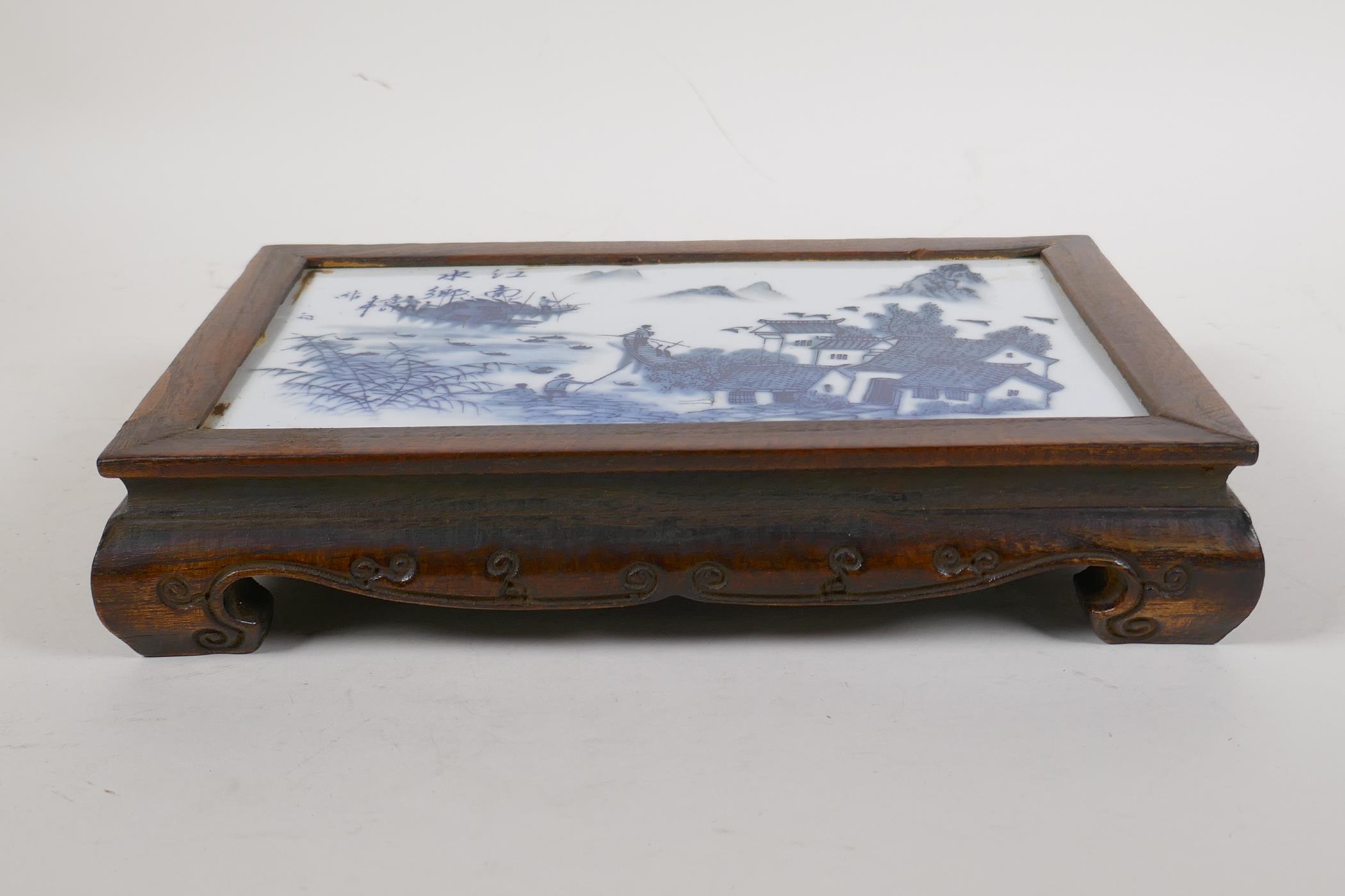A Chinese hardwood stand mounted with a blue and white porcelain panel depicting a riverside