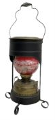 Brass oil lamp with red painted glass shade encased in black metal with handle and four feet