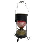 Brass oil lamp with red painted glass shade encased in black metal with handle and four feet
