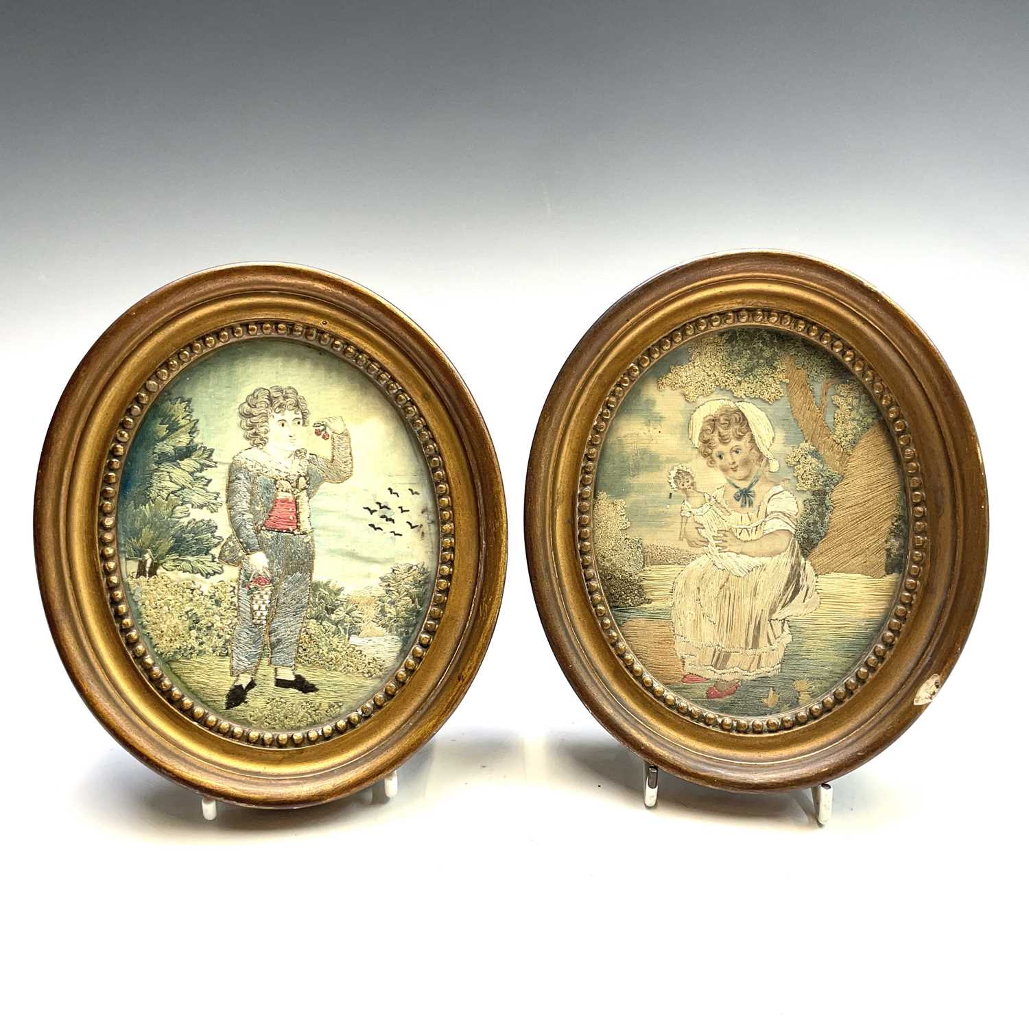 A pair of Regency small oval silk work pictures, with a boy picking fruit and a girl with a doll,