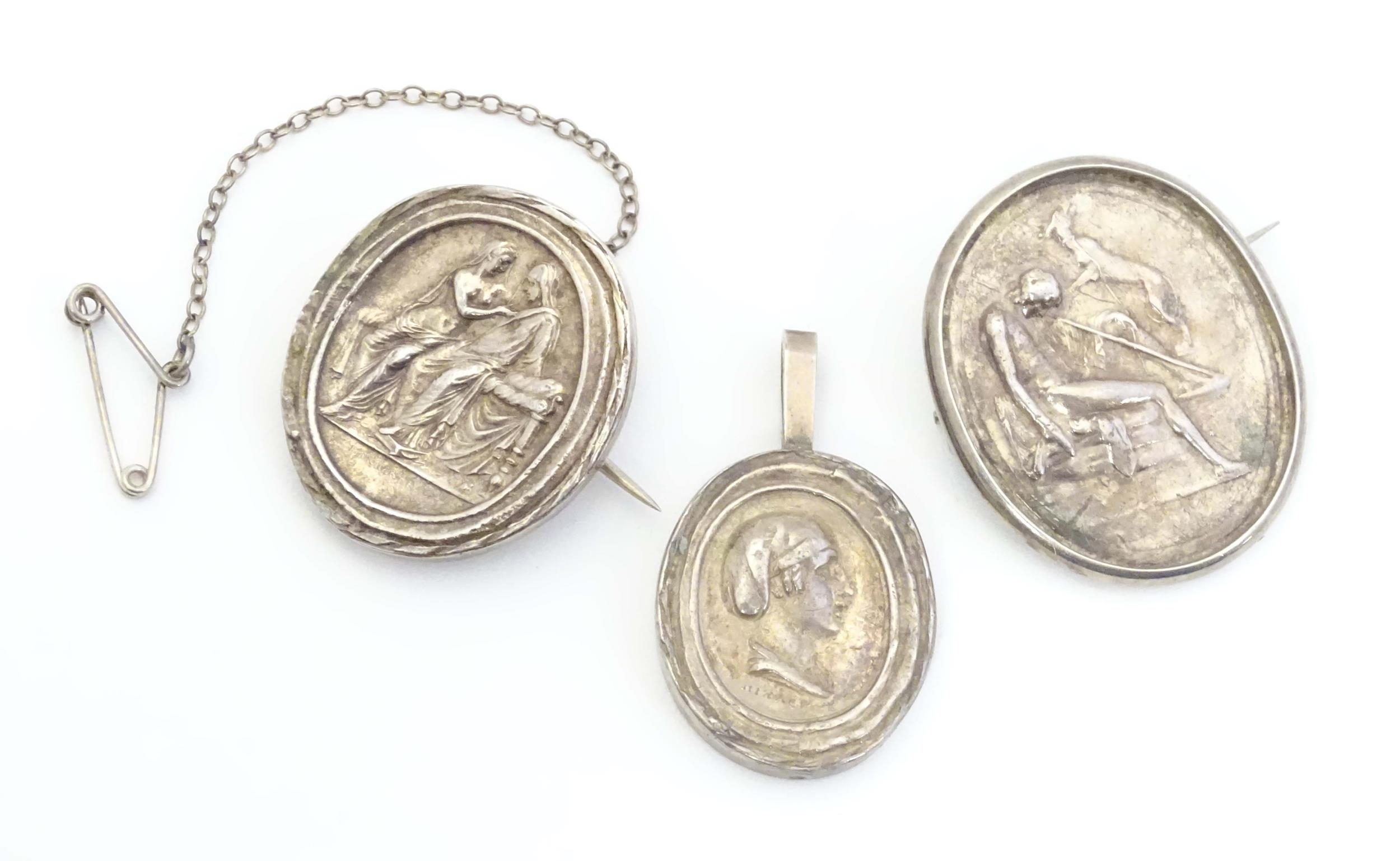 Two white metal brooches of oval form set with various classical scenes together with a white