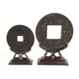 Two oversized cast replicas of Chinese cash coins on carved wooden stands, one decorated with a