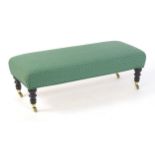 A long upholstered stool raised on four turned tapering legs terminating in brass caps and