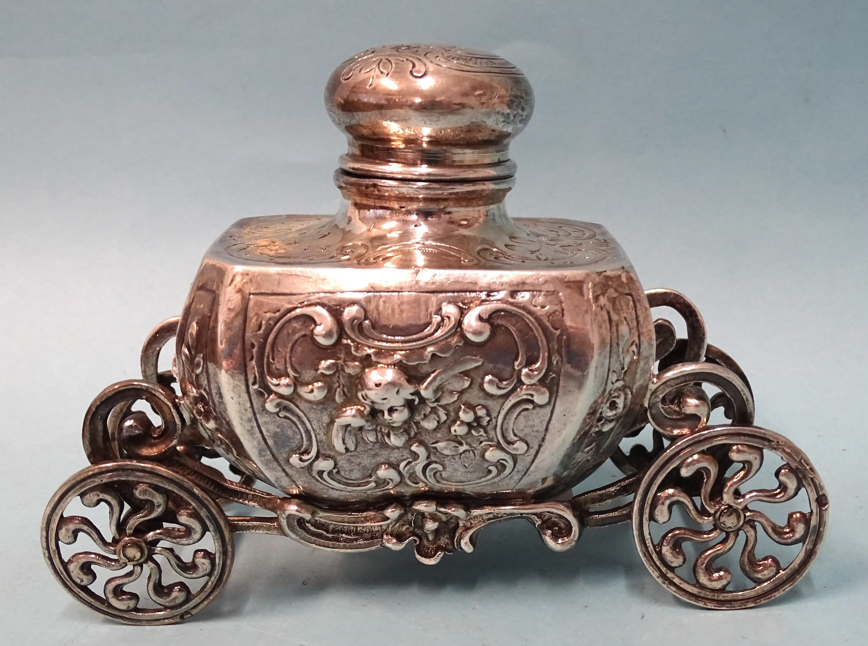 A silver inkwell of Baroque form, raised on ornate carriage with four wheels, 9cm high, 12.5cm long,