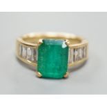 A modern yellow metal and single stone emerald set dress ring, with graduated baguette cut diamond